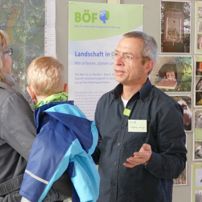 Ein Experte erklärt einer jungen Mutter mit Kind ein Projekt vor einer Fotostellwand.