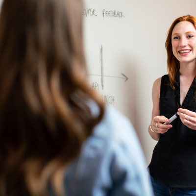 Junge Frau steht mit einem Stift an einem Whiteboard und lächelt eine weitere Frau mit langen Haaren an, die man von hinten sieht.