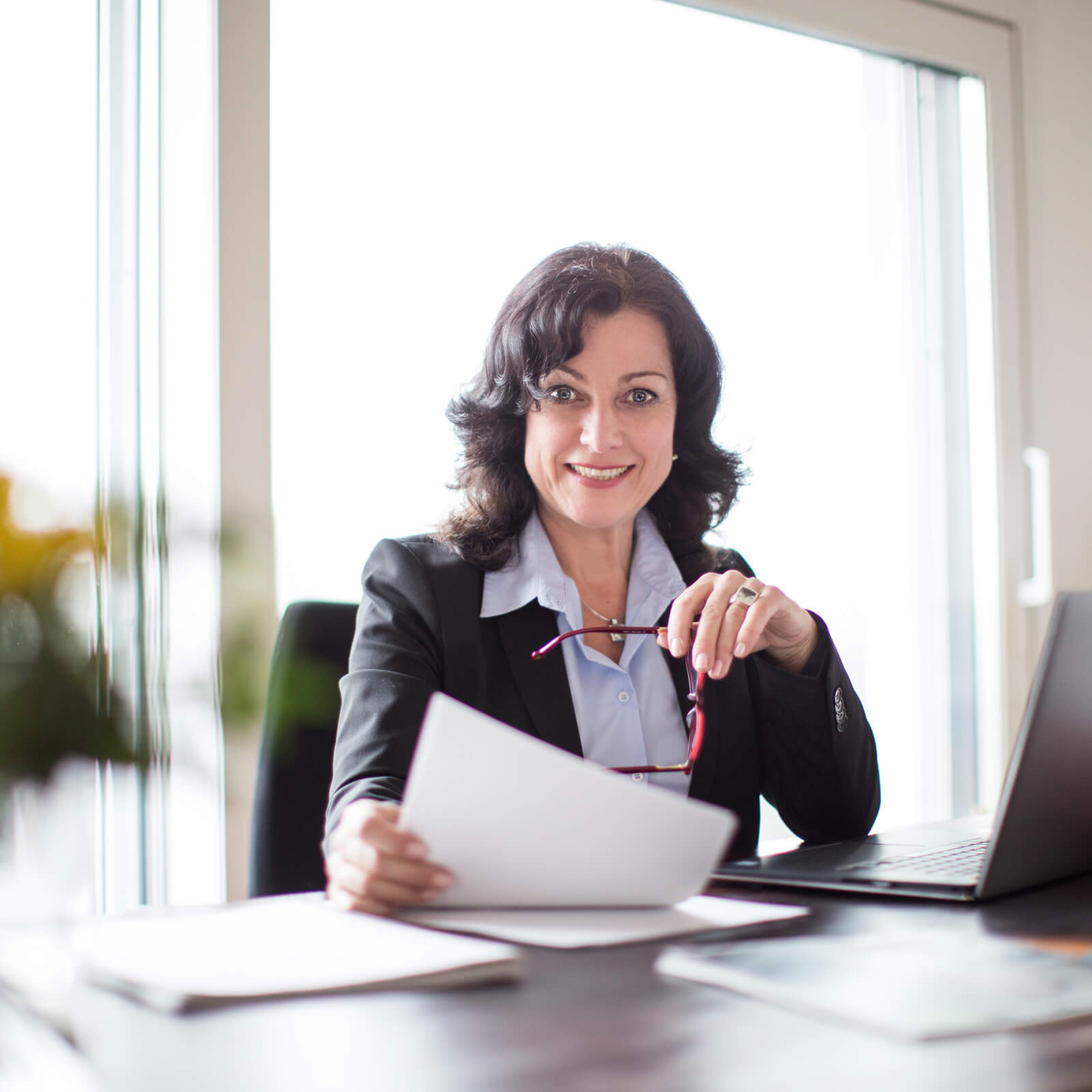 Frau in Businesskleidung sitzt an Schreibtisch vor einem Notebook, sie hält Papiere und eine Brille in den Händen und lächelt in die Kamera.