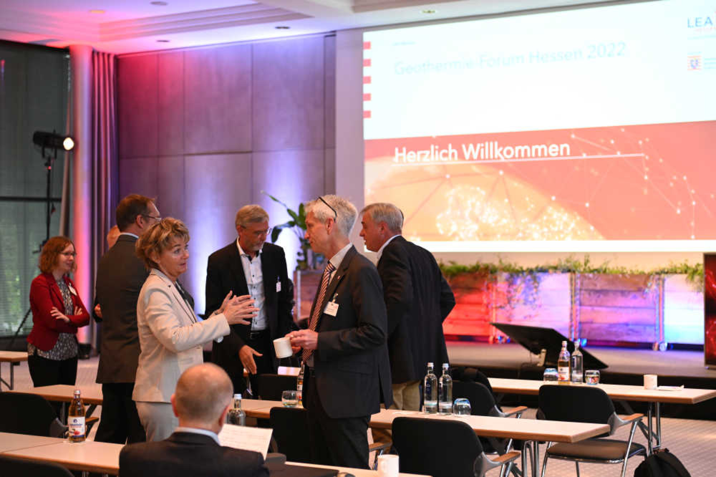 Geothermie-Forum 2022: Bühne des Vortragssaals Bühne eines Vortragssaals mit Großbildschirm und einer Gruppe Menschen, die zwischen Tischreihen steht und sich unterhält.