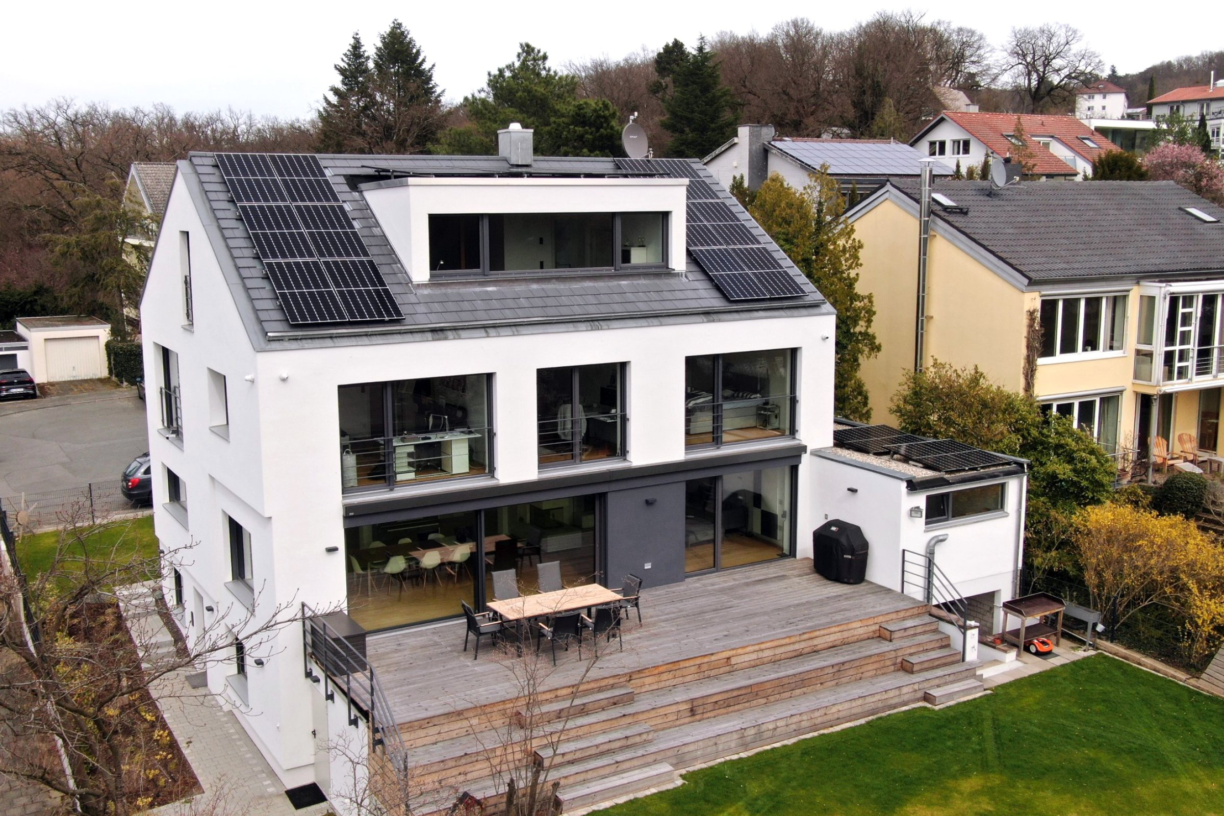 Rückansicht des Hauses von Familie Bednarek in Darmstadt mit großen Fensterflächen und Photovoltaik auf dem Dach sowie einer mehrstufigen Holzterasse zum Garten.