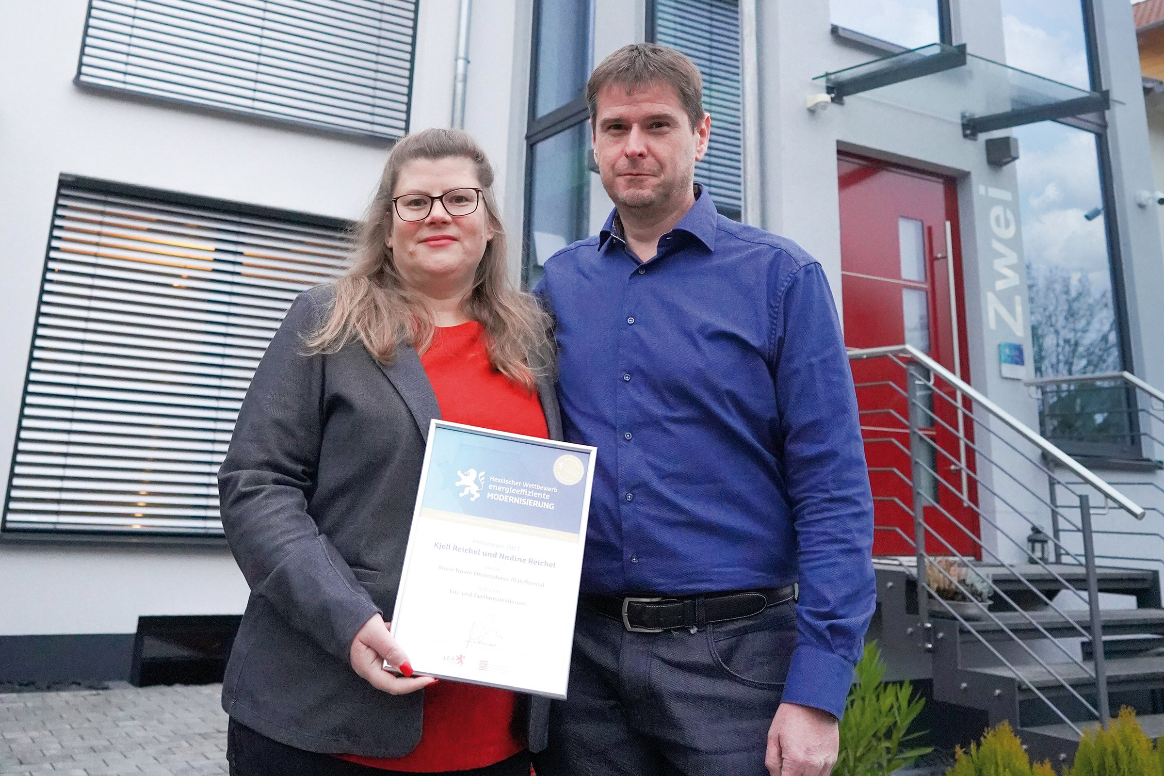 Ehepaar Reichel steht mit einer Urkunde vor ihrem Haus mit roter Tür.