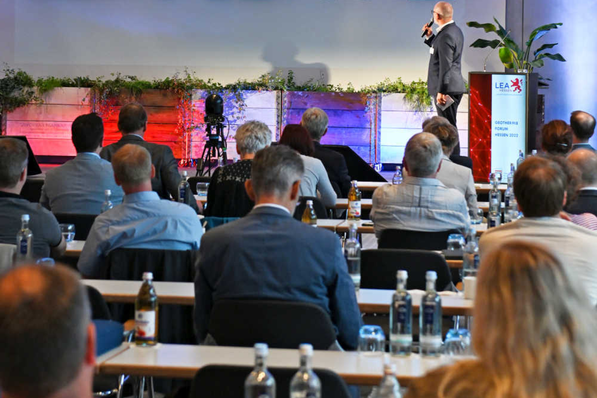 Geothermie-Forum 2022: Gespräch mit Onlineteilnehmern am Bildschirm Mann mit Mikrophon steht auf einer Bühne vor einem geteilten Großbildschirm, auf dem zwei weitere Männer zu sehen sind. Im Raum sitzen Zuschauer an langen Tischreihen.