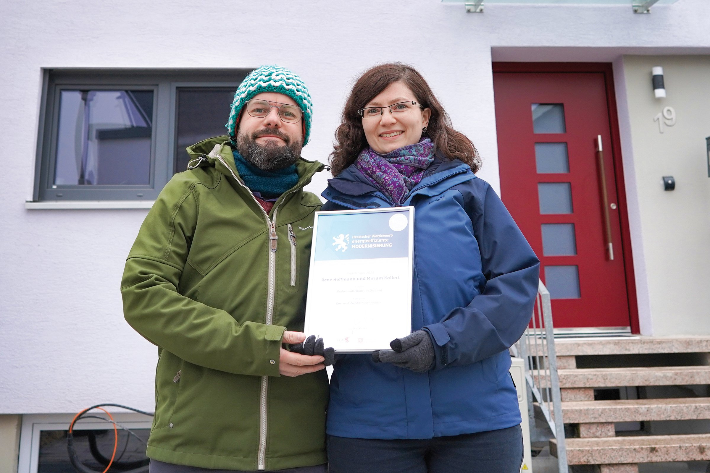 René Hoffmann und Miriam Kallert stehen mit der Urkunde für den 2. Preis vor ihrem Haus in Winterkleidung.
