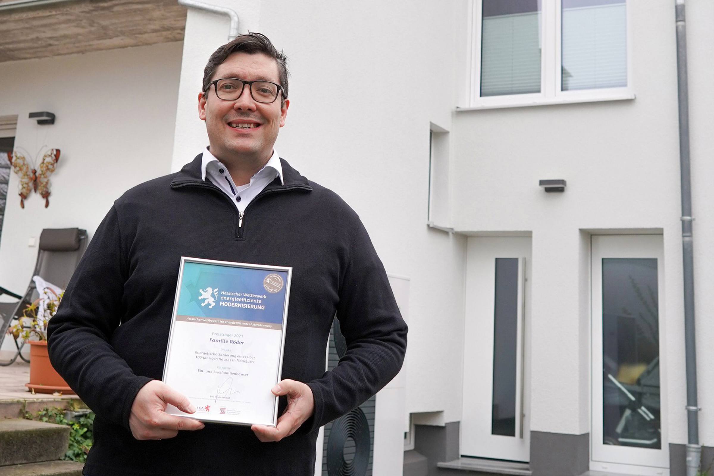 Herr Röder steht mit der Urkunde für den 3. Preis vor der Rückseite seines Hauses.