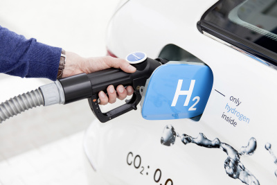 Hand hält einen Tankstutzen in die Tanköffnung eines weißen PKW. Auf dem Tankdeckel steht "H2".