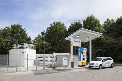 Weißer PKW steht an einer überdachten Wasserstofftankstelle mit Bäumen im Hintergrund.