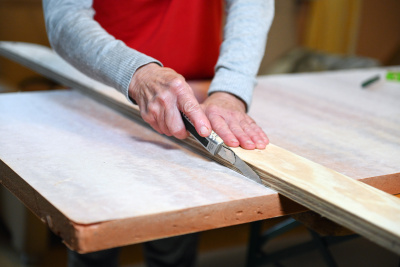 Ältere Frau schneidet Dämmmaterial mit einem Messer an einer Messlatte aus Holz entlang.