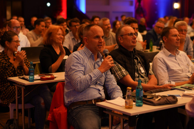 Mann im Zuschauerraum sitzt mit Mikrofon vor seinem Tisch und stellt eine Frage.