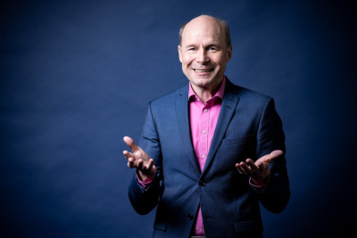 Porträt von Karsten McGovern, Geschäftsführer der LEA Hessen. Er steht mit ausgebreiteten Händen vor einem dunklen Hintergrund in Anzug mit pinkfarbenen Hemd.