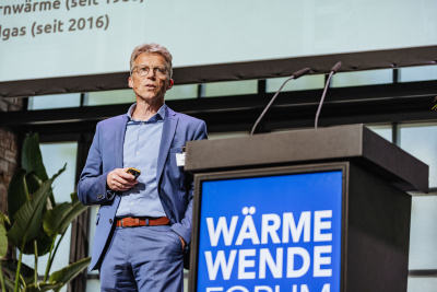 Wärmewende Forum Hessen: Vortragender Markus Hardt steht auf der Bühne neben dem Renderpult mit einer Fernbedienung in der Hand.