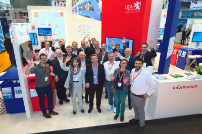 Gruppe von Menschen steht an einem Messestand mit roter Säule, auf der das LEA-Logo mit Löwenfigur zu sehen ist. Alle winken in die Kamera.