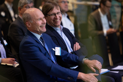 LEA-Geschäftsführer Dr. Carsten McGovern sitzt im Publikum bei einem Vortrag. Er und ein Mann neben ihm lachen.