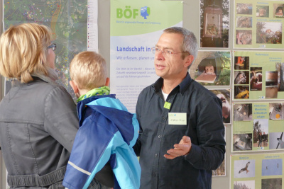 Ein Experte erklärt einer jungen Mutter mit Kind ein Projekt vor einer Fotostellwand.