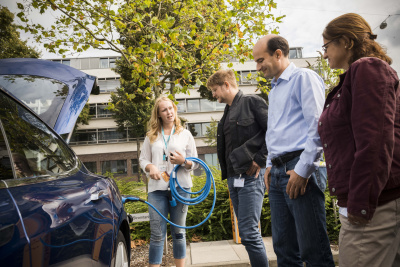 Seminarleiterin erklärt das Laden eines E-Autos mit einem Stromstecker einer Gruppe von Menschen, die vor einem blauen Auto steht.