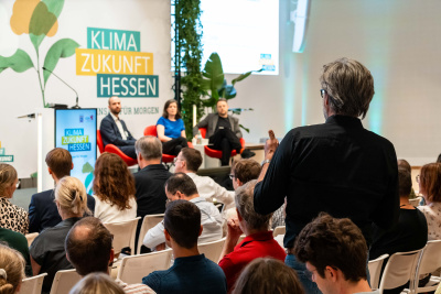 Moderator und Referentinnen auf der Bühne vor vielen Menschen im Publikum, ein Mensch aus dem Publikum stellt eine Frage