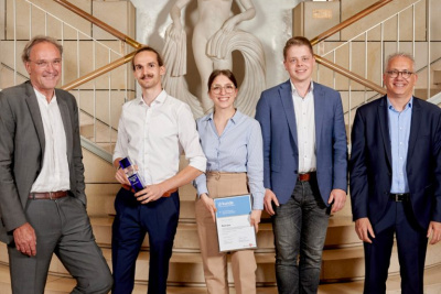 Gruppe von vier Personen steht vor einer Foyer-Treppe, zwei Personen halten eine Auszeichnung in den Händen.