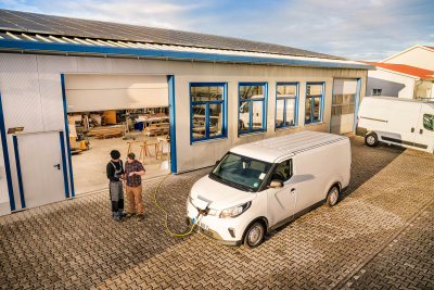 Photovoltaik-Anlage auf dem Dach einer Lagerhalle eines KMU. Vor der Lagerhalle steht ein Elektroauto, das l&auml;dt und zwei Mitarbeiter des Unternehmens, die auf ein Tablet schauen.