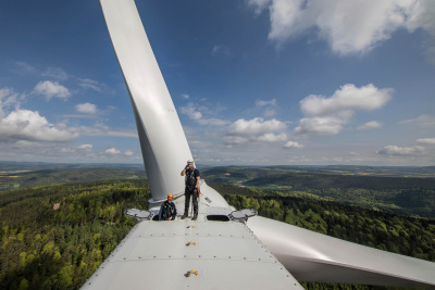 Zwei Wartungsarbeiter auf einem Windrad, im Hintergrund eine hügelige Waldlandschaft.