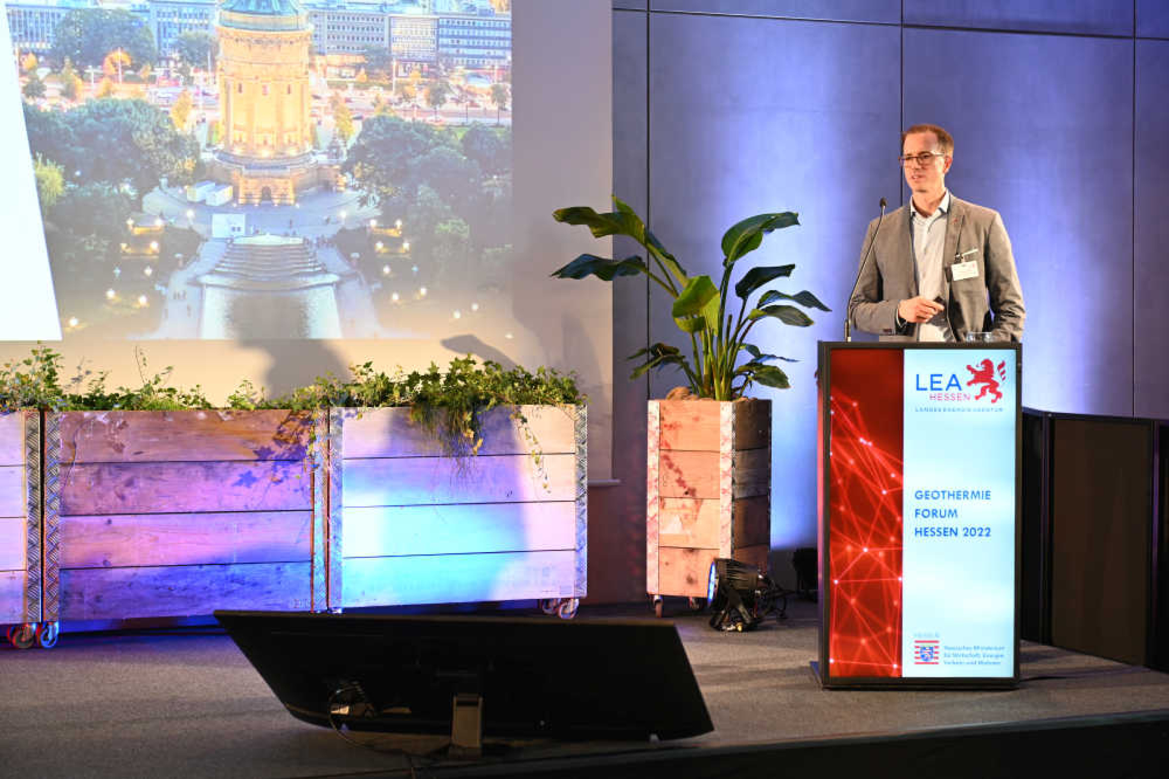 Geothermie-Forum 2022: Vortrag Dr. Kristian Bär Ein Mann steht an einem Rednerpult mit der Aufschrift "LEA – Geothermie Forum Hessen 2022", neben ihm auf einem Großbildschirm sieht man ein erleuchtetes Gebäude.