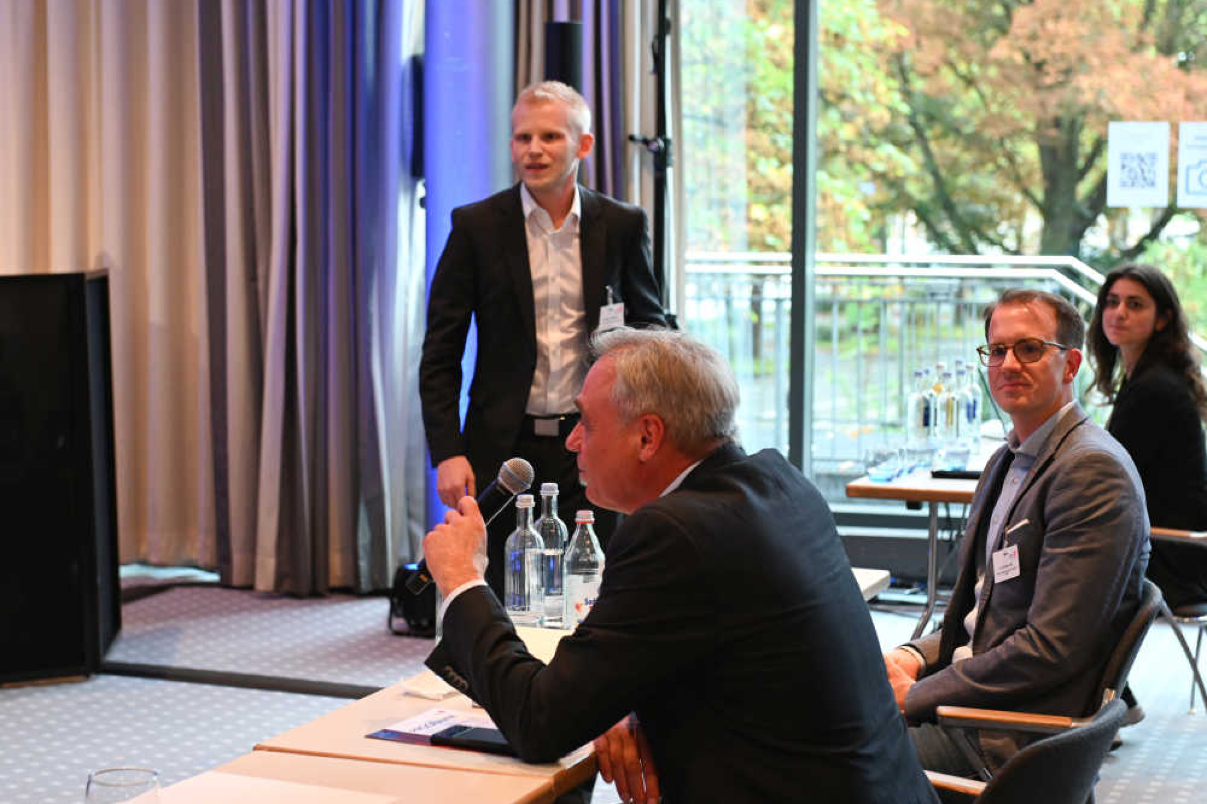 Geothermie-Forum 2022: Fragen aus dem Zuschauerraum In einem größeren Raum sitzen Männer und Frauen an längeren Tischen. Ein Mann hält ein Mikrofon in der Hand und spricht. Im Hintergrund steht ein weiterer Mann vor einer Fensterwand mit Ausblick auf herbstliche Bäume.