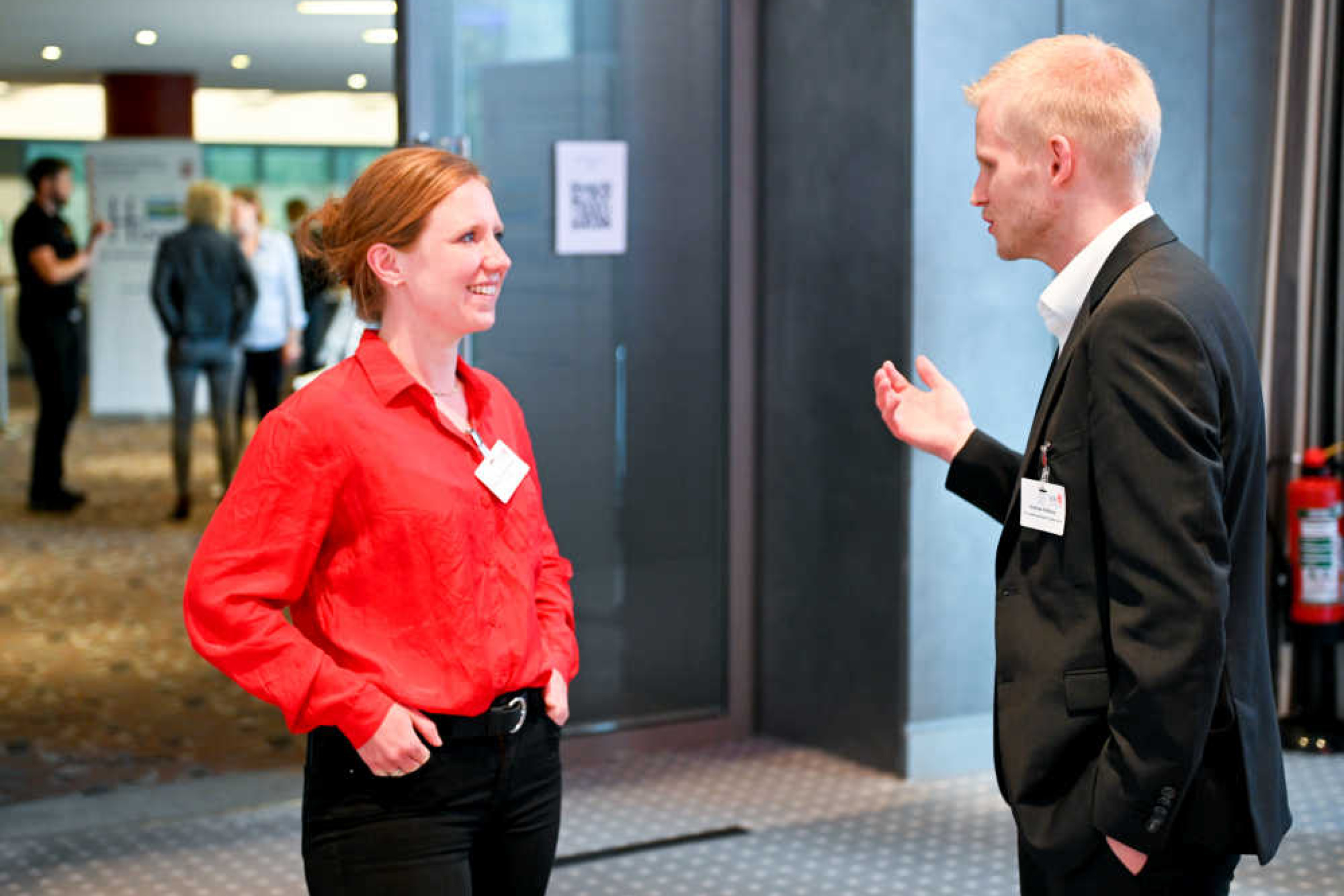 Geothermie-Forum 2022: Gespräch in der Pause Frau und Mann stehen in einem Raum und sprechen miteinander. Im Hintergrund sieht man den Übergang zu einem weiteren Raum, in dem eine Gruppe von Personen zusammensteht.