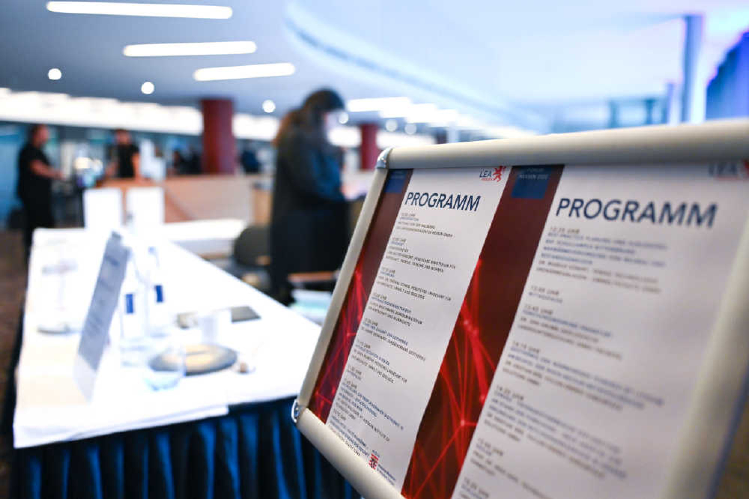 Geothermie-Forum 2022: Stellwand Programm Stellwand in einem großen Raum mit zwei Blättern, auf denen "Programm" steht. Im Hintergrund lange Tische mit Infomaterial und Menschen.