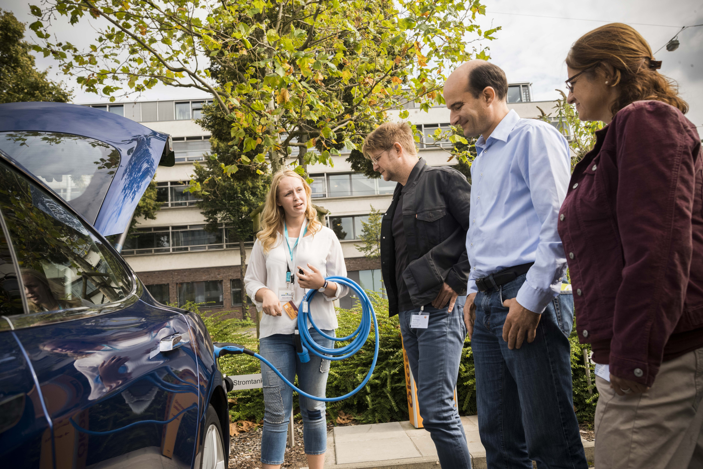 Seminarleiterin erklärt das Laden eines E-Autos mit einem Stromstecker einer Gruppe von Menschen, die vor einem blauen Auto steht.