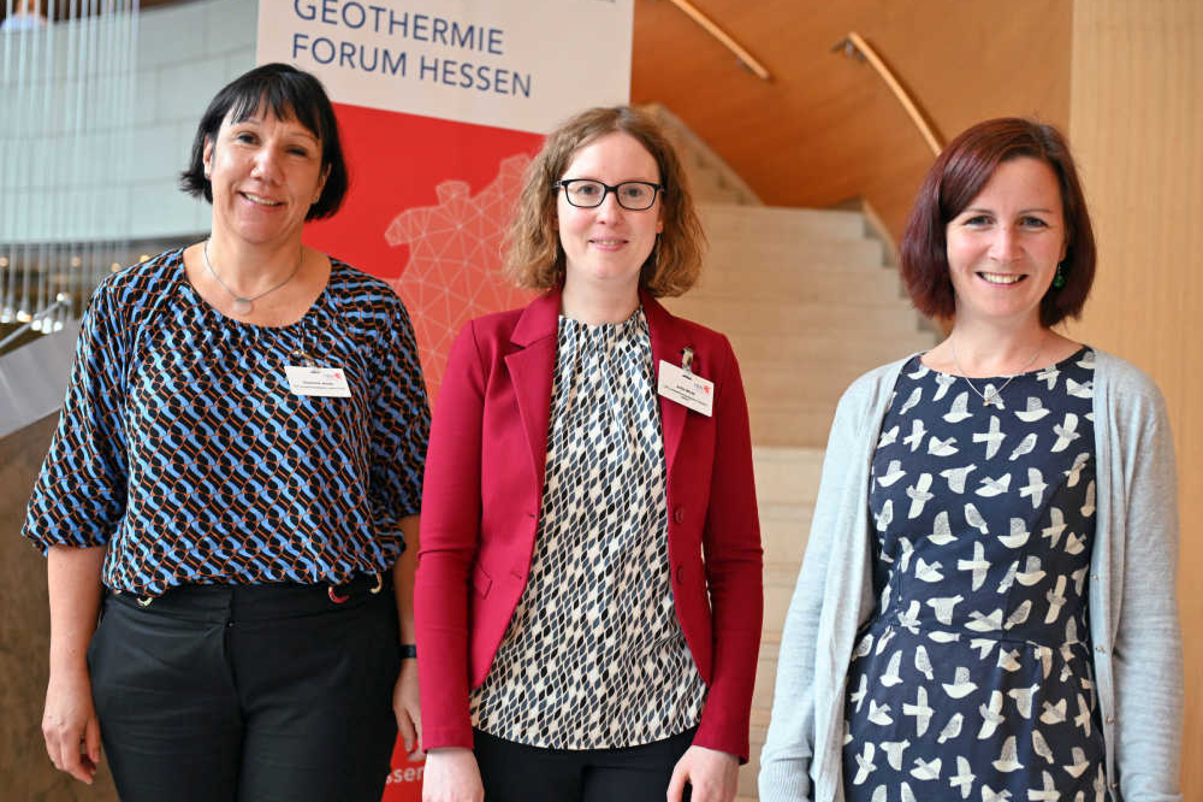 Drei Frauen stehen nebeneinander vor einer Treppe und lächeln. Hinter ihnen ist eine Stellwand mit der Aufschrift "Geothermie Forum Hessen".