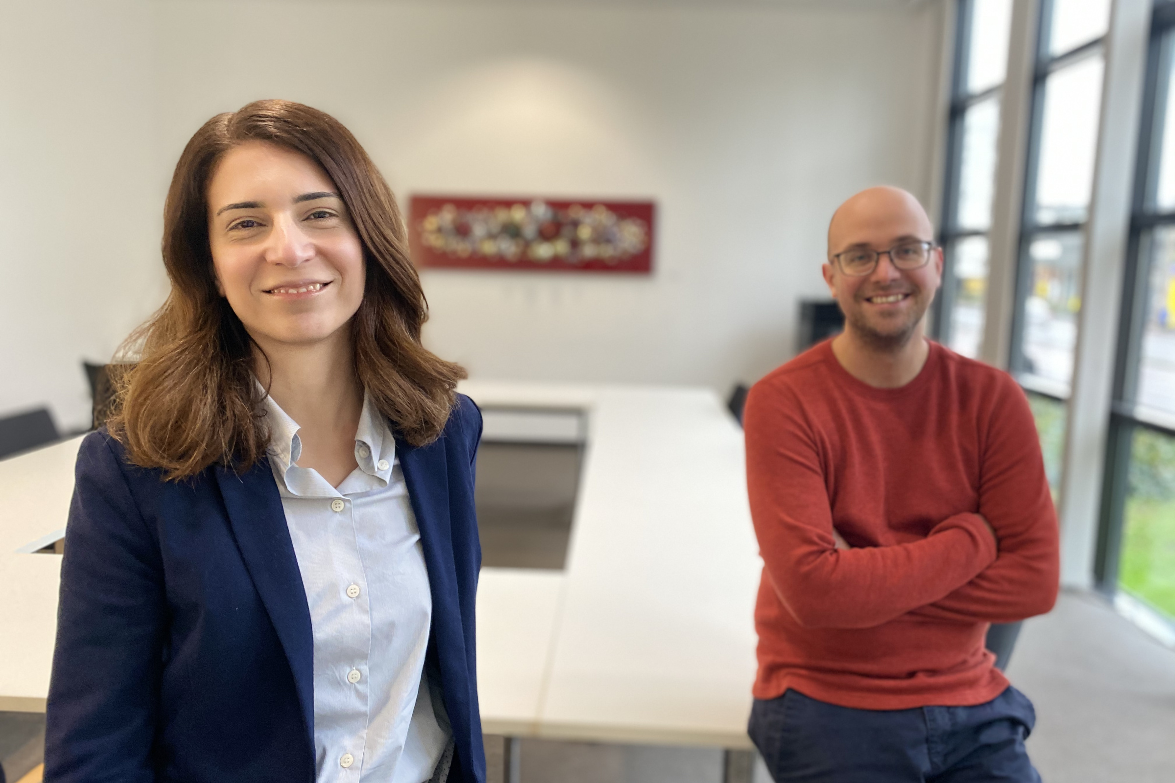 Eine Frau und ein Mann in einem leeren Konferenzraum lächeln in die Kamera.