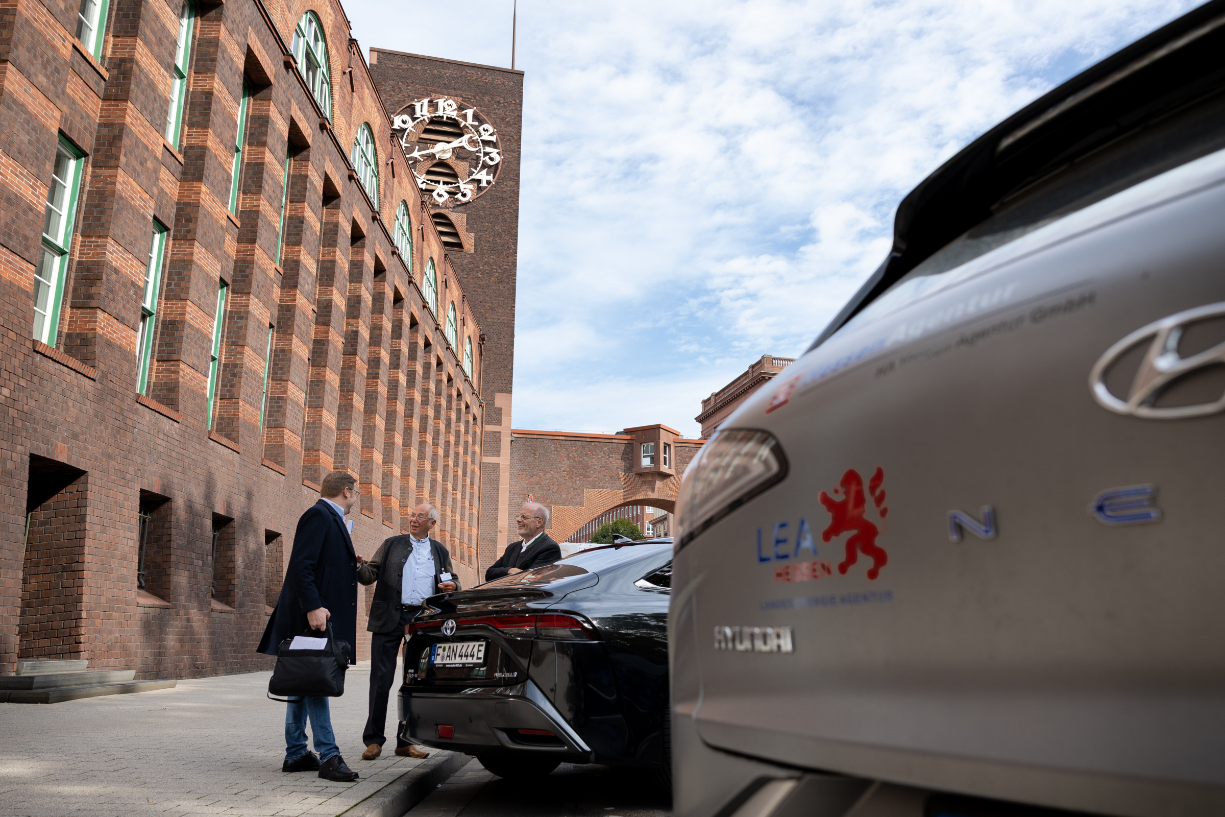 Brennstoffzellenforum 2022 Mehrere Männer stehen vor einem Gebäude, vor dem Autos parken. Im Vordergrund ein PKW mit dem Logo der LEA Hessen.