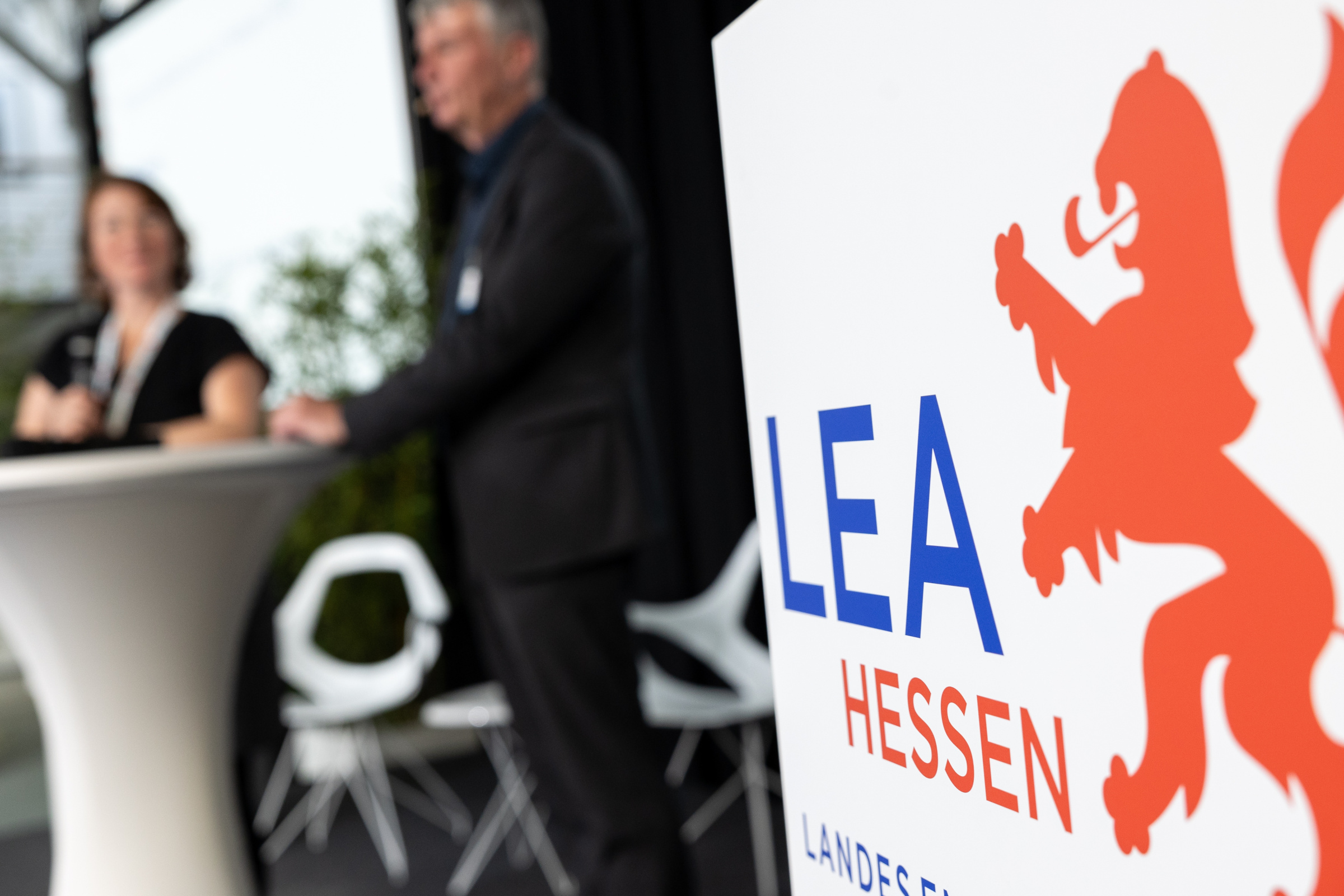 Logo der Lea Hessen an einer Stellwand, im Hintergrund zwei Personen an einem weißen Stehtisch.
