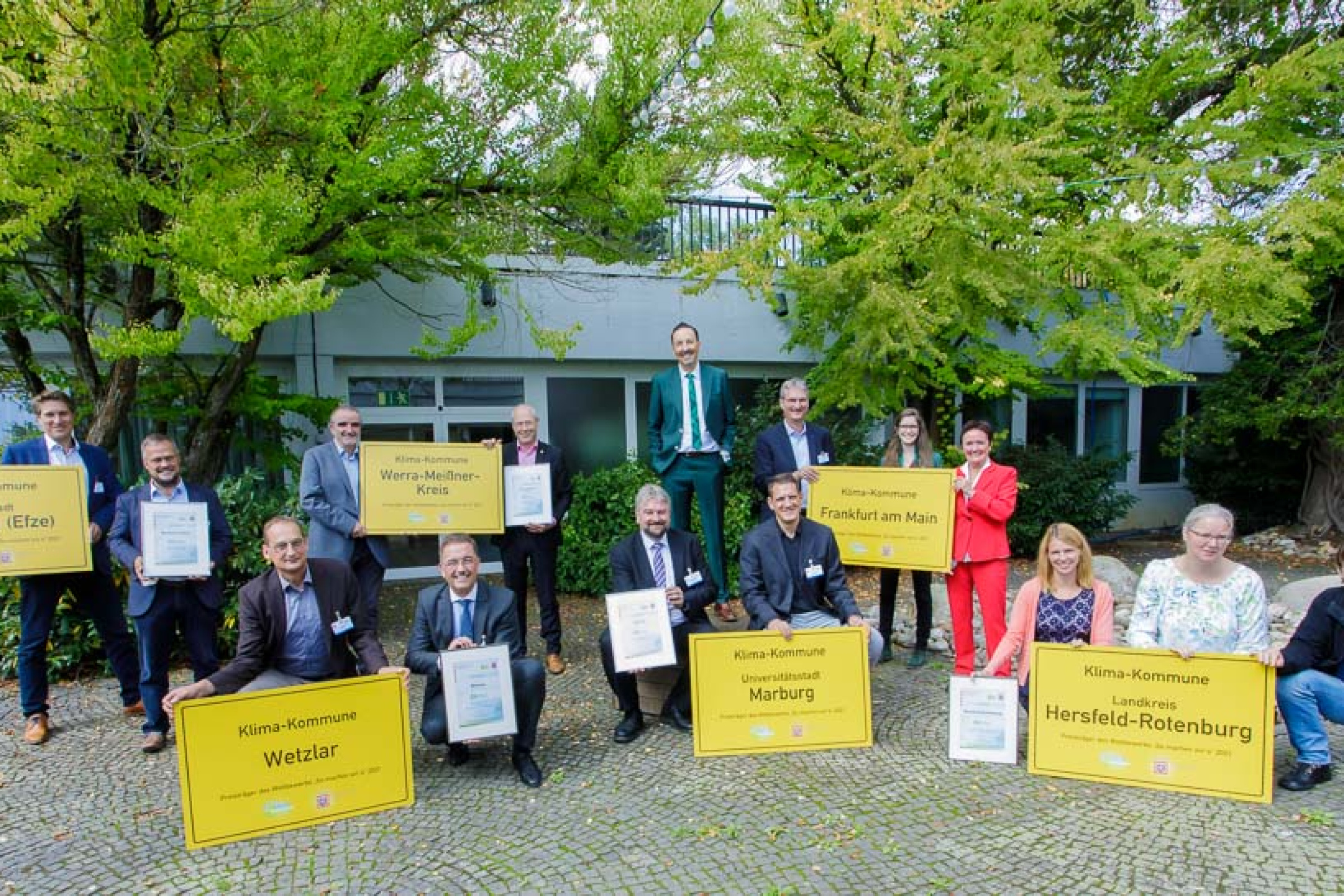 Klima Kommunal 2021: Gruppe von Menschen hocken und stehen mit gelben Ortsschildern vor einem Gebäude.