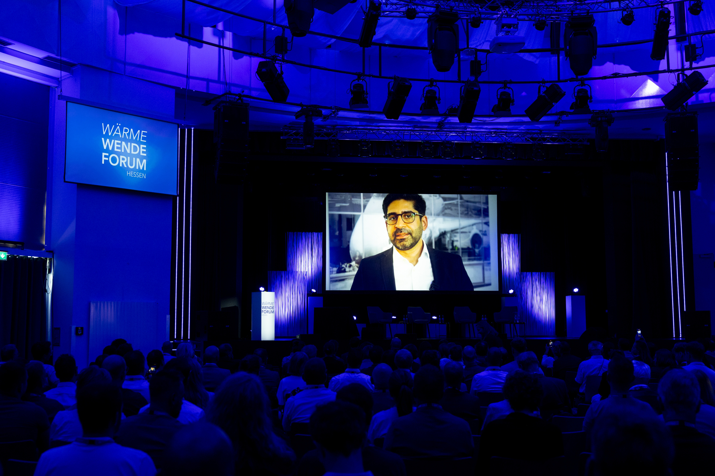 Großer Saal mit vielen Menschen, auf einer Leinwand wird eine Videobotschaft gezeigt