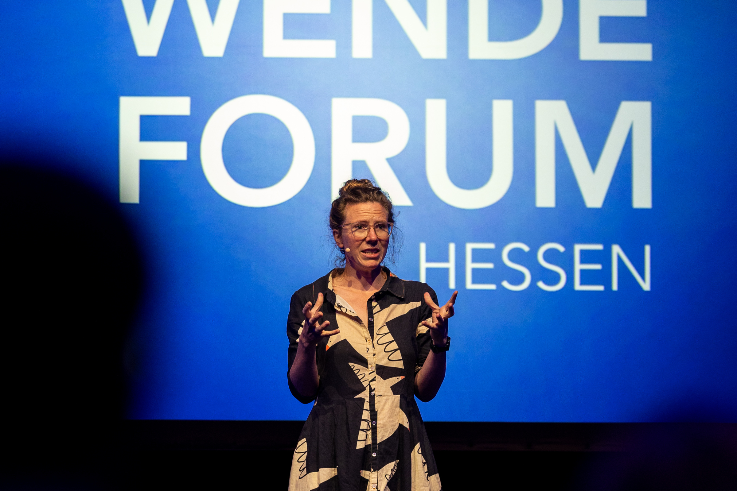 Eine Frau auf der Bühne vor einem Mikro, im Hintergrund ein Bildschirm mit dem Logo des Wärmewende Forum Hessen