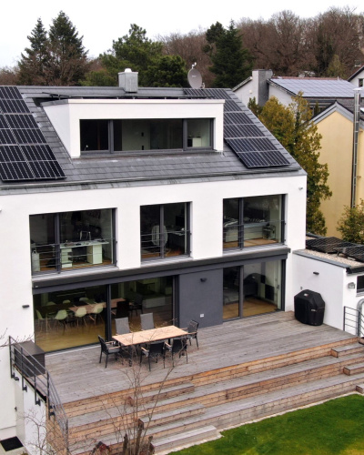 Rückansicht des Hauses von Familie Bednarek in Darmstadt mit großen Fensterflächen und Photovoltaik auf dem Dach sowie einer mehrstufigen Holzterasse zum Garten.
