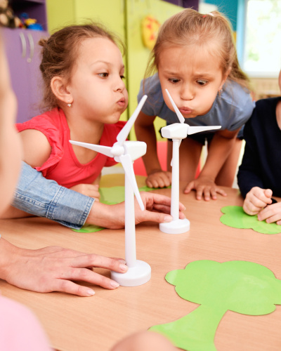 Einige Kindergartenkinder sitzen an einem Tisch und pusten auf Windrad-Modelle.