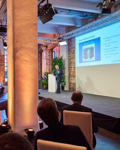 Gesamtansicht Veranstaltung Brennstoffzellenforum mit Videoleinwand und Zuschauern auf Sesseln.