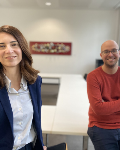 Eine Frau und ein Mann in einem leeren Konferenzraum lächeln in die Kamera.