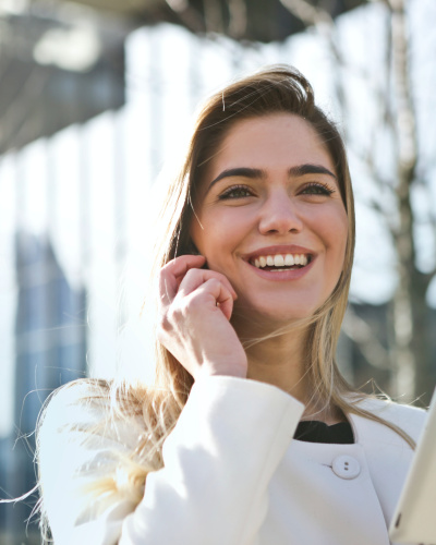 Junge Frau mit langen blonden Haaren in heller Jacke steht im Freien vor einem Gebäude, telefoniert lächelnd und hält in der anderen Hand ein Tablet.