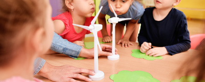 Einige Kindergartenkinder sitzen an einem Tisch und pusten auf Windrad-Modelle.