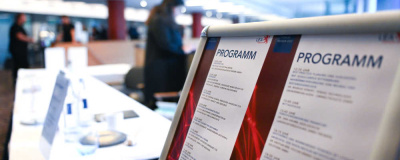 Geothermie-Forum 2022: Stellwand Programm Stellwand in einem großen Raum mit zwei Blättern, auf denen "Programm" steht. Im Hintergrund lange Tische mit Infomaterial und Menschen.