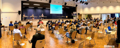 Saal mit Zuschauern beim Bürgerforum Energiewende Hessen. Auf der Bühne sitzen einige Personen an Tischen mit einer großen Videoleinwand im Hintergrund.