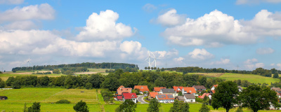 Naturlandschaft mit Feldern und Wald, im Vordergrund ein Dorf, im Hintergrund mehrere Windr&auml;der.