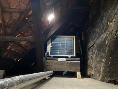 Die Außeneinheit einer Luft-Wasser-Wärmepumpe steht auf dem durchlüfteten Dachboden eines Fachwerkhauses.