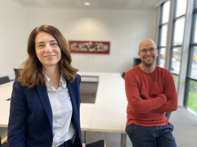 Eine Frau und ein Mann in einem leeren Konferenzraum lächeln in die Kamera.