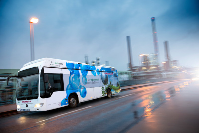 Wasserstoff-Bus der Firma Winzenhöler fährt auf einer Straße, im Hintergrund ein Kraftwerk oder eine Raffinerie.