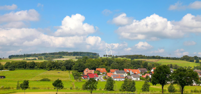 Naturlandschaft mit Feldern und Wald, im Vordergrund ein Dorf, im Hintergrund mehrere Windräder.